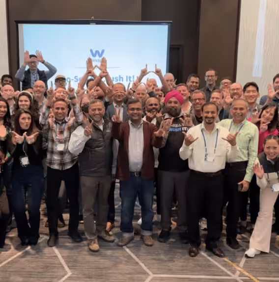 Group of diverse people posing indoors, many making hand gestures and smiling toward the camera.