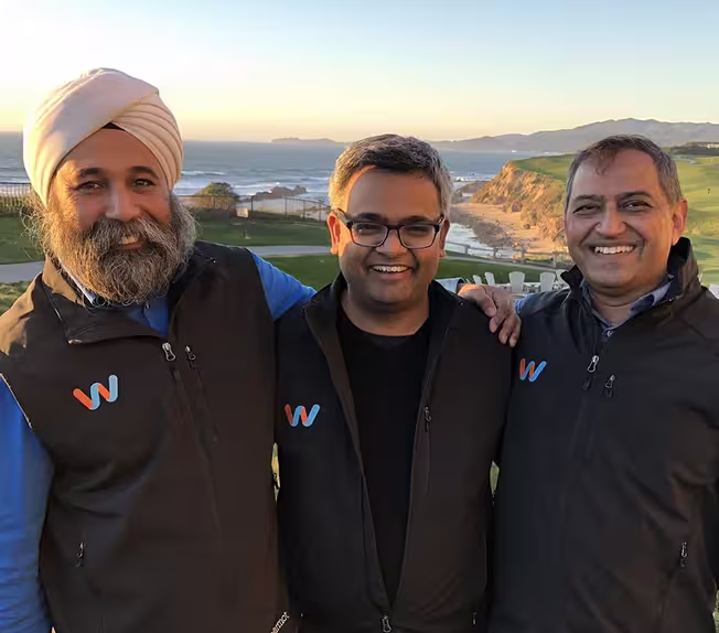 Three men smiling outdoors wearing black vests with a W logo, scenic ocean and cliffs in the background.